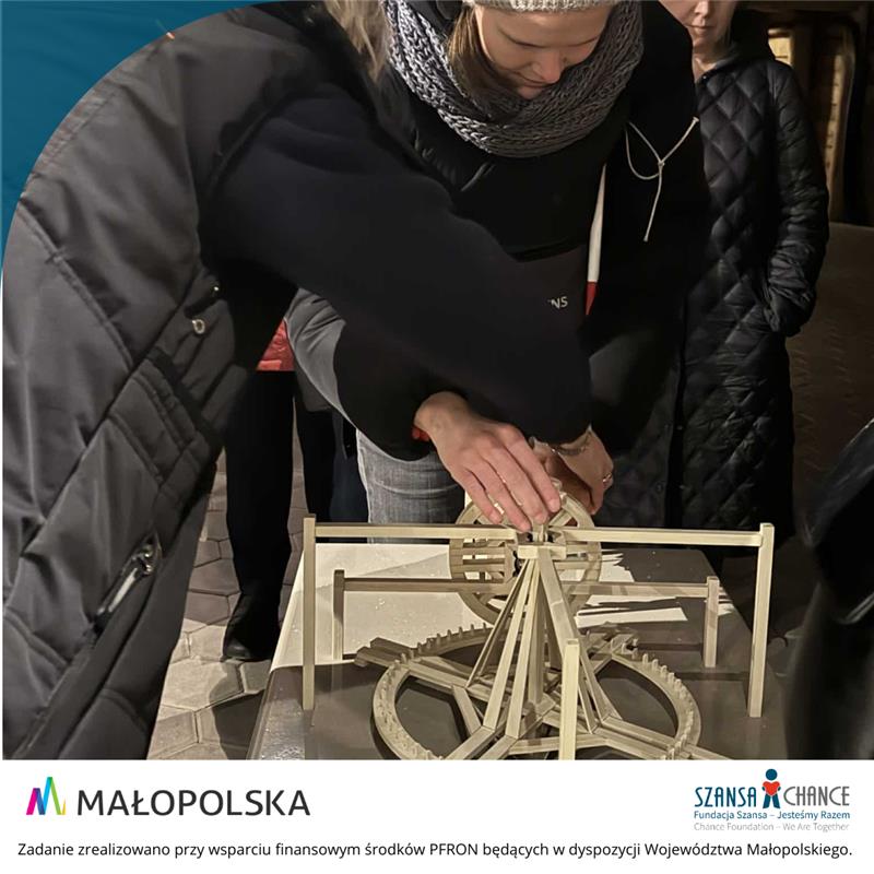 Zdjęcie przedstawia ręce pani przewodnik oraz ręce dziewczyny, która dotyka pomniejszonej makiety kieratu, przy którym pracowały konie w kopalni.